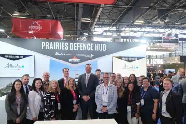 A large group of people wearing business attire and lanyards pose for a photo. Behind them is a sign that reads Prairies Defence Hub.