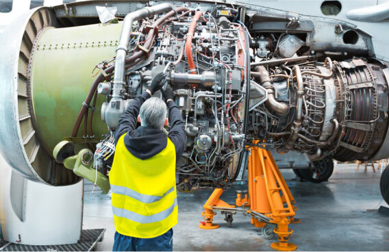 A technician in a safety vest works on an exposed aircraft engine in a hangar.