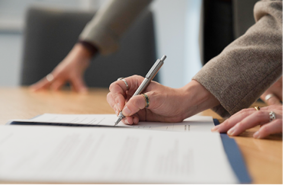 Person signing document on wooden table in formal meeting setting.