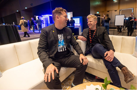 Two professionals seated on couch discussing at corporate networking event with booths in background.