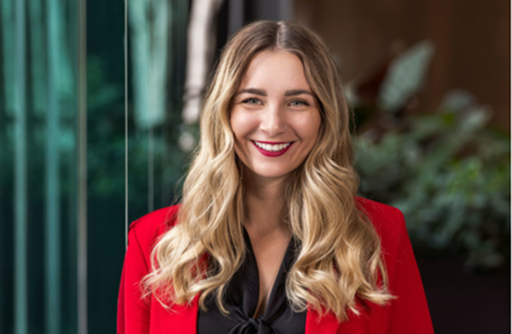 Woman with long blonde hair wearing red blazer and black blouse standing indoors near glass wall with natural light.