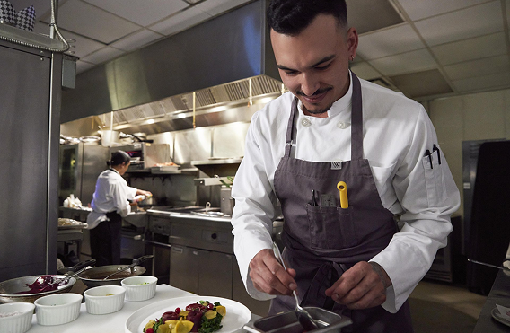 Chef plating colorful dish with sauce while another works near stove.