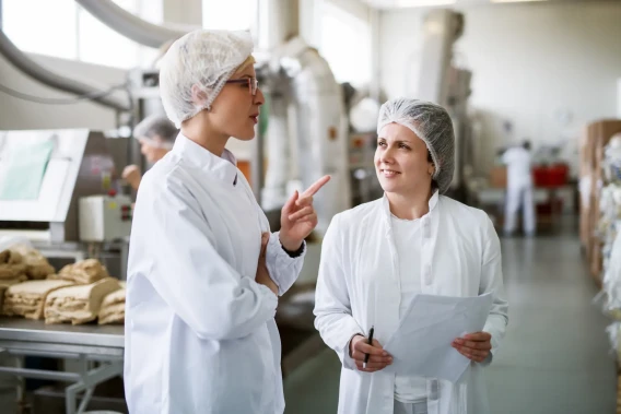 Two workers in lab coats and hairnets talk inside a food processing facility.