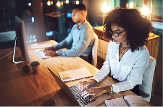 Two people working at desk with laptop and desktop computer under warm lights.
