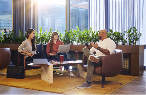 Three colleagues collaborating with laptops and smartphone in modern lounge with plants and natural light.