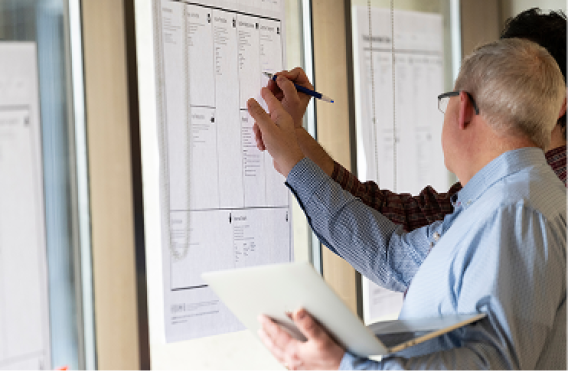 Two professionals examining printed charts taped to glass with laptop and pen.