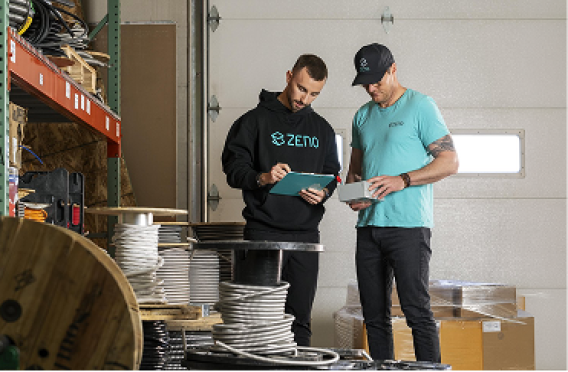Two workers in warehouse with branded clothing reviewing notes and tablet among spools and boxes.