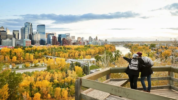 Two people overlooking the Calgary skyline and river valley filled with autumn trees from a wooden viewpoint.