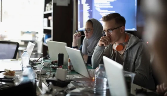 Developers working on laptops in a shared office with code displayed on a screen in the background.