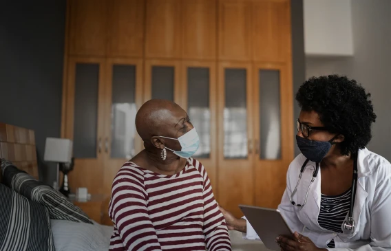 Doctor wearing a mask consulting with a patient at home while holding a tablet.
