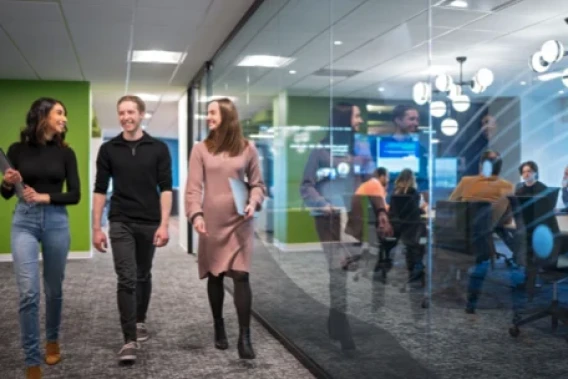 Three employees walking through a modern office hallway beside a glass-walled meeting room.
