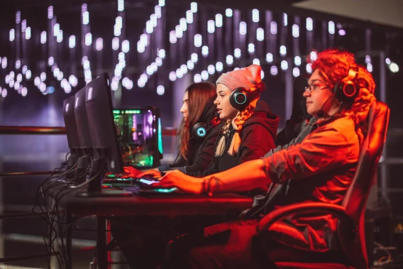 Three gamers wearing headsets compete at desktop computers in a neon-lit gaming event.