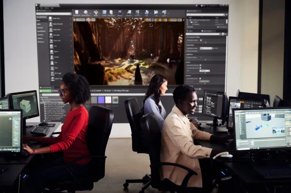 Three people work at desktop computers in a game development lab with a large projected Unreal Engine screen behind them.