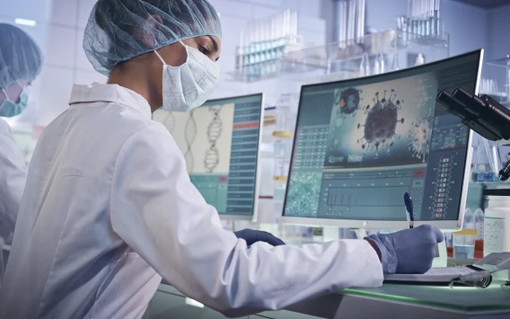 Masked researcher analyzing viral and genomic data on laboratory computer monitors.
