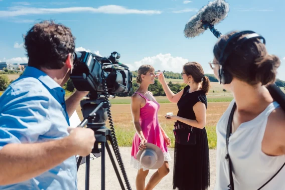 Outdoor film shoot with a camera operator, makeup artist, boom microphone, and on-camera talent in a sunny field.