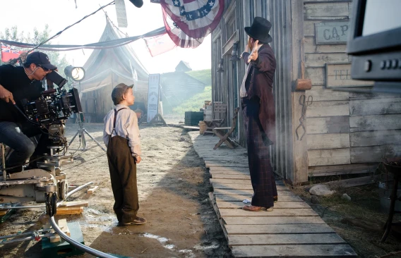 Camera crew filming a scene with actors on a period-style outdoor movie set.