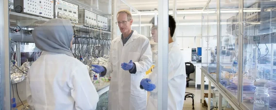 Three scientists in lab coats discussing work inside a bright research laboratory.