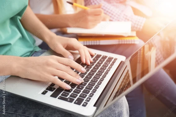 Close-up of hands typing on a laptop while another person writes in a notebook nearby.