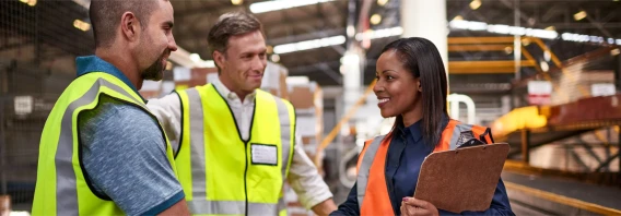 Warehouse staff in high-visibility vests having a discussion on the warehouse floor, with one holding a clipboard.