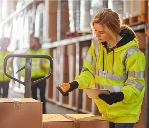 Warehouse worker in high-visibility clothing scanning packages while holding a tablet in a distribution center.