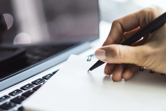 Close-up of a hand writing notes in a notebook beside a laptop keyboard.