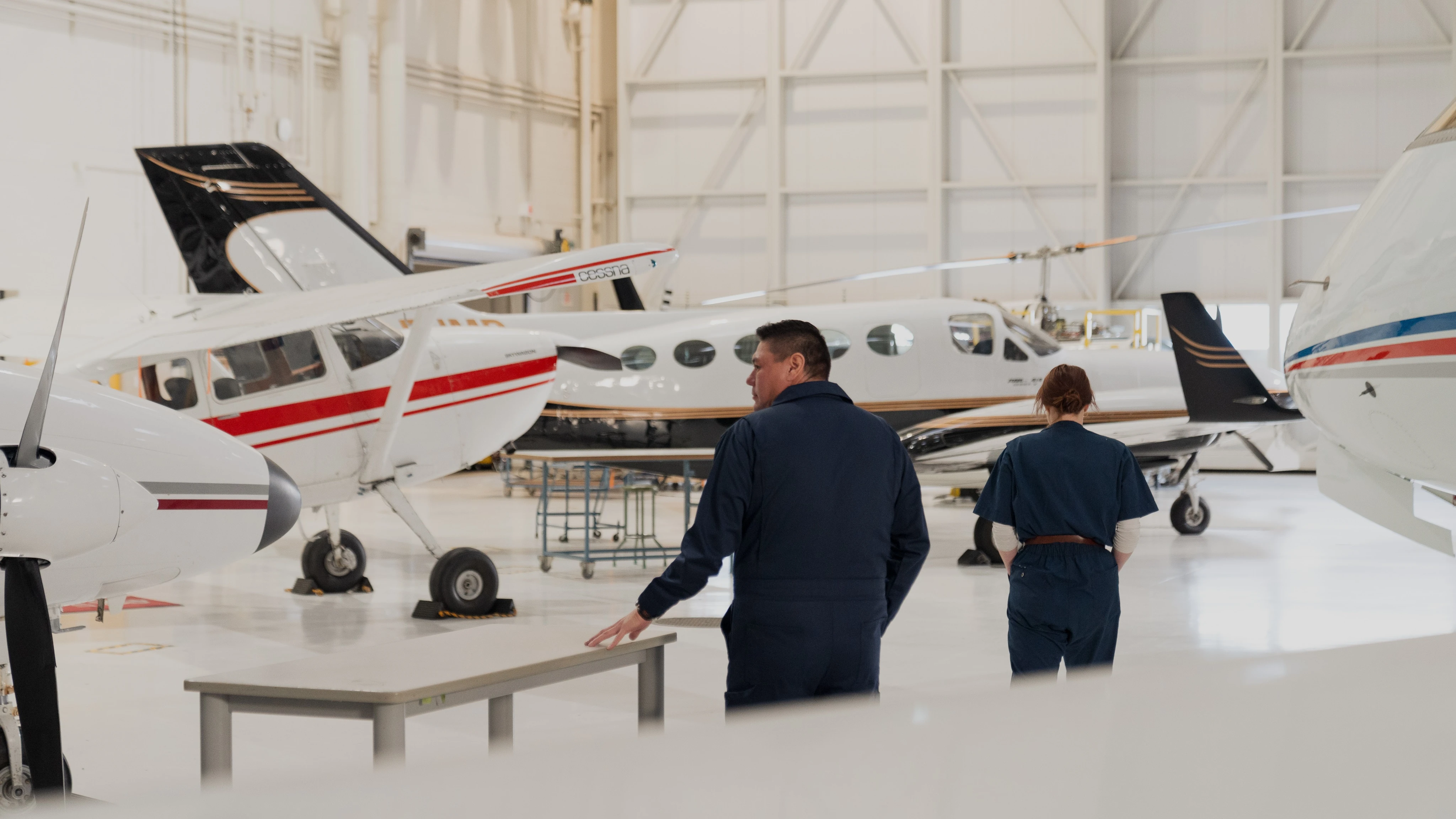 - Technicians in coveralls inspecting small airplanes inside spacious, well-lit hangar.