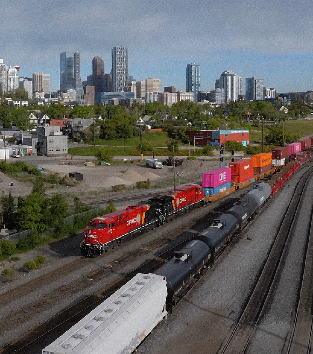 Red locomotives pulling freight cars in train yard with city skyline in background.