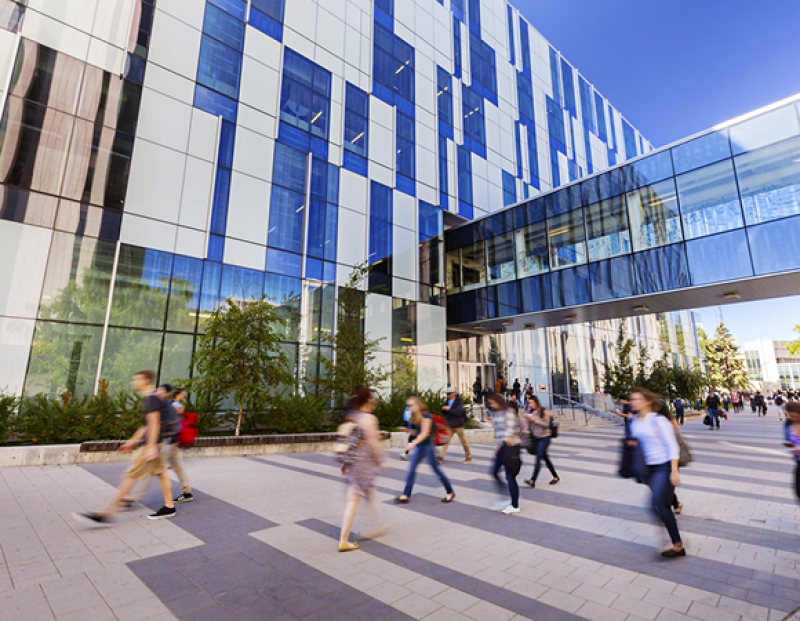 People walking outside a modern glass building with a skybridge connecting two sections.