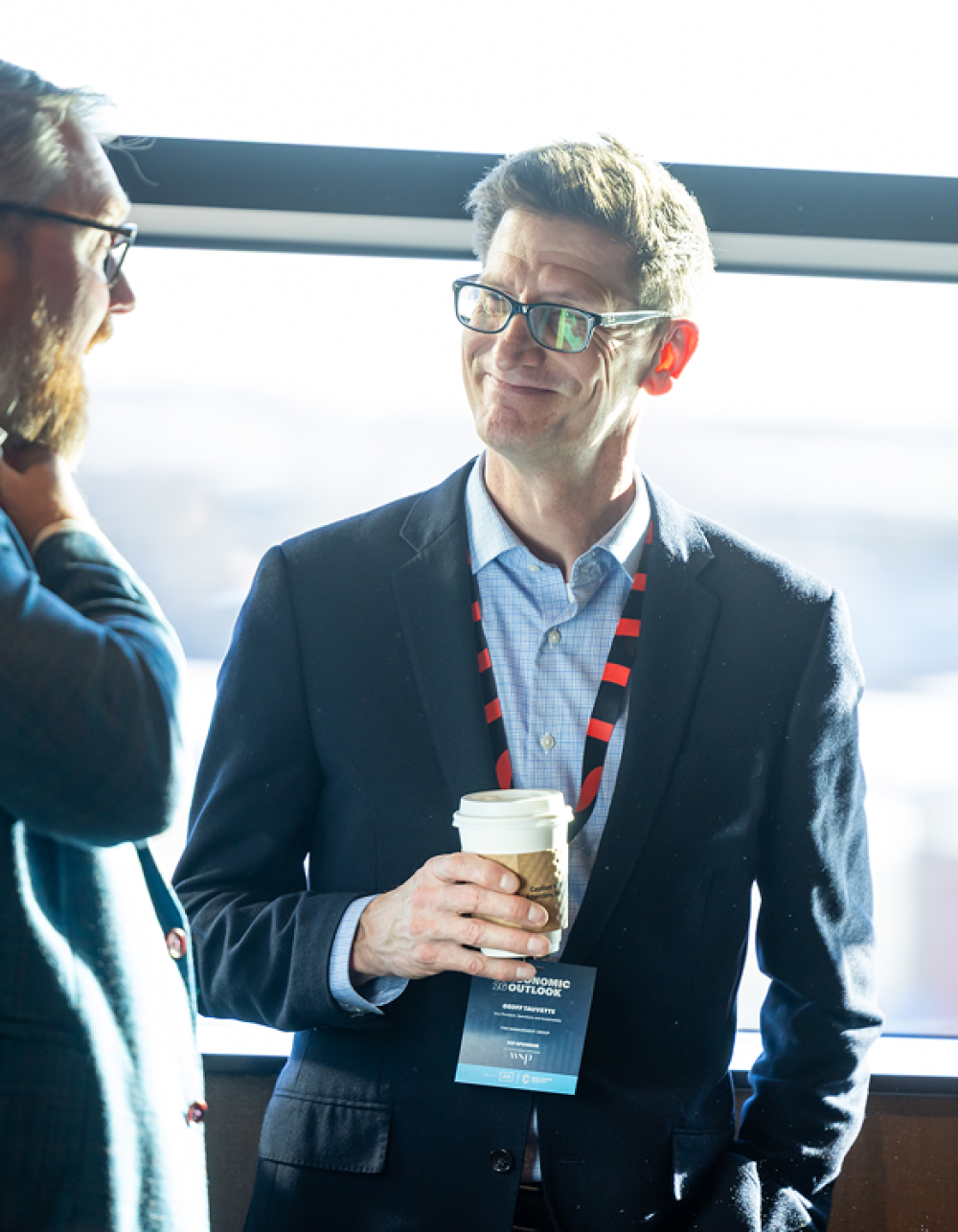 Two professionals talking at economic outlook event, one wearing VIP badge and holding coffee cup