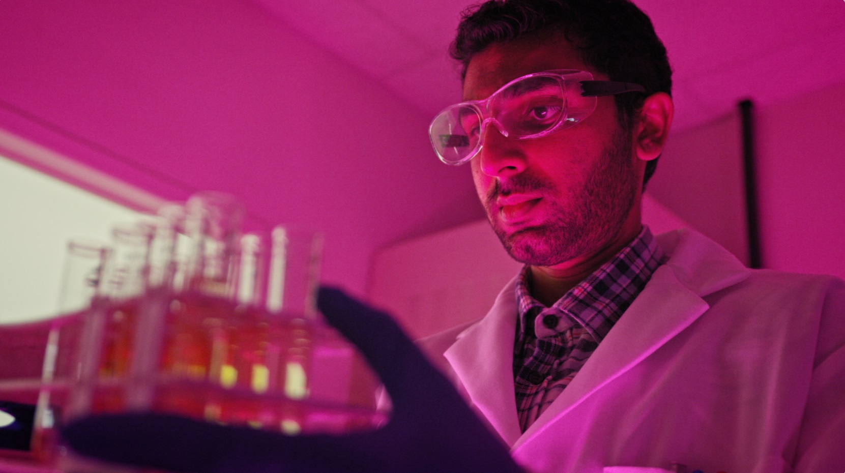 Researcher in lab coat holding rack of colorful test tubes under dramatic pink lighting.