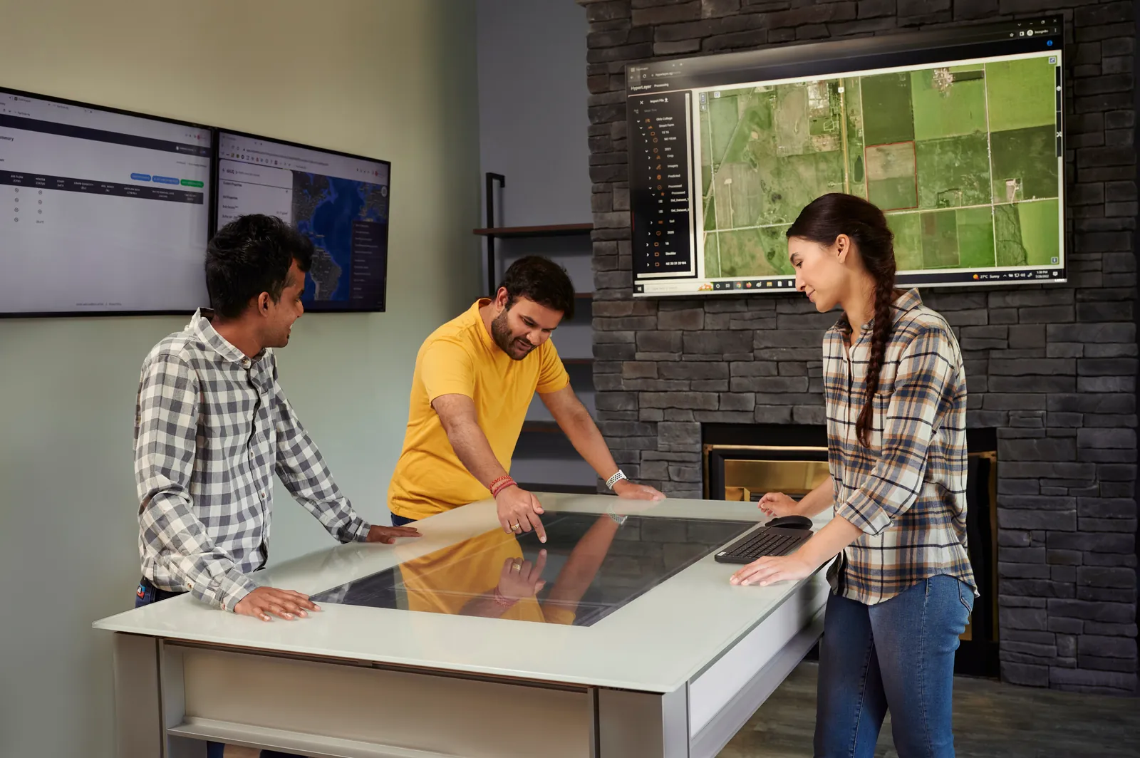Three people reviewing farm data on a touchscreen table with maps displayed on wall monitors.