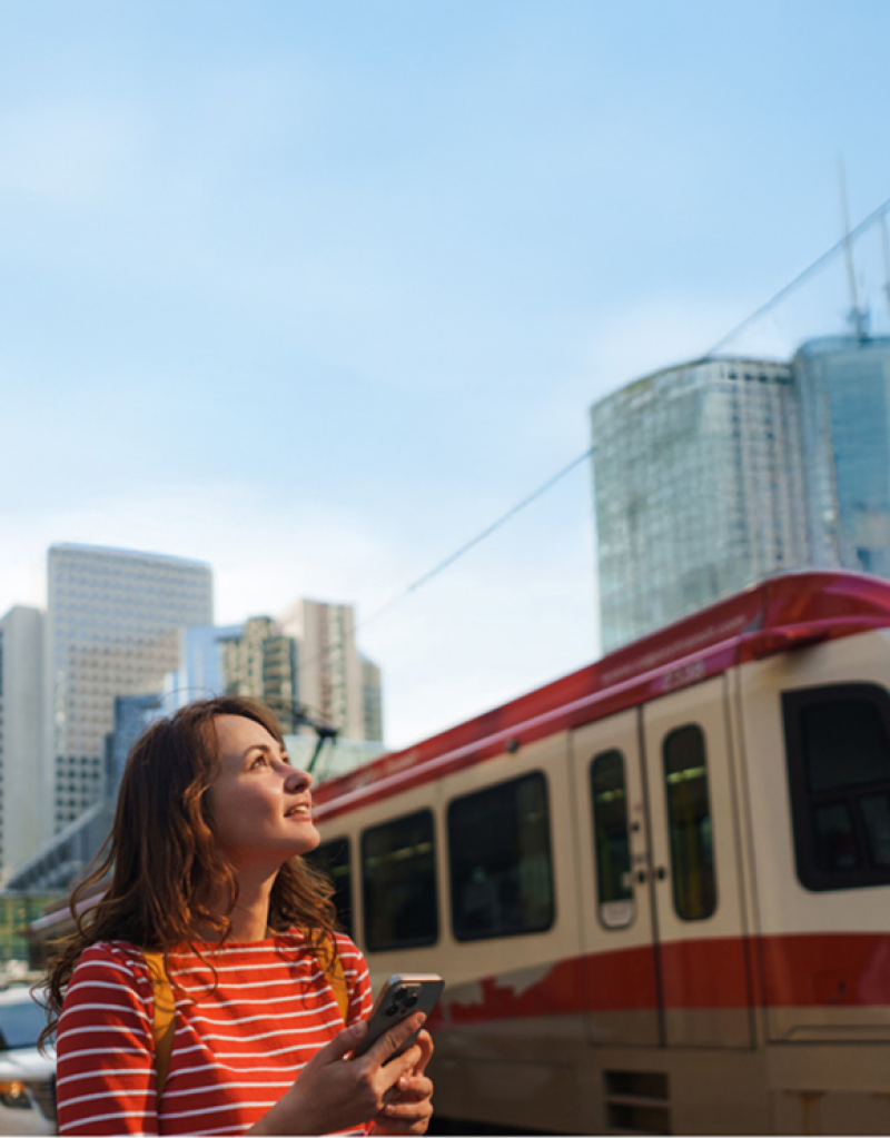 Person with backpack holding phone near light rail and tall buildings.