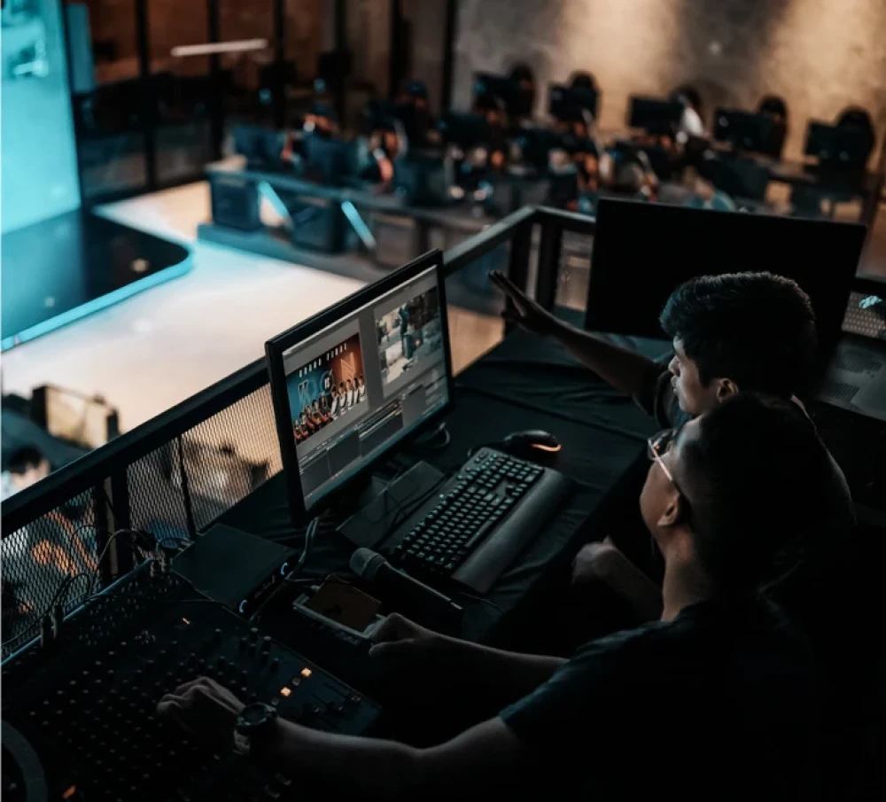 Two people operate video and audio equipment from a control booth overlooking a stage.