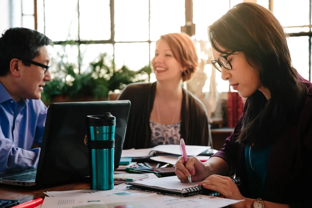 Three students studying together at a table with notebooks, a laptop, and coffee mugs.